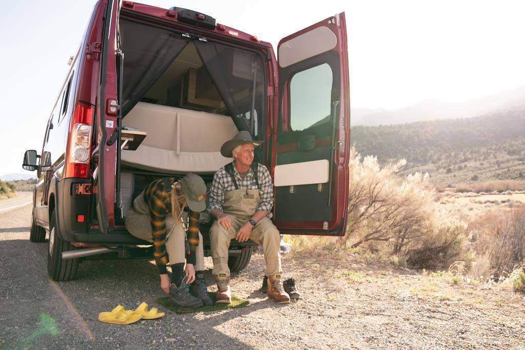 A photo of an older man and younger woman putting on boots to go fly-fishing while seated in the back of their camper van.
