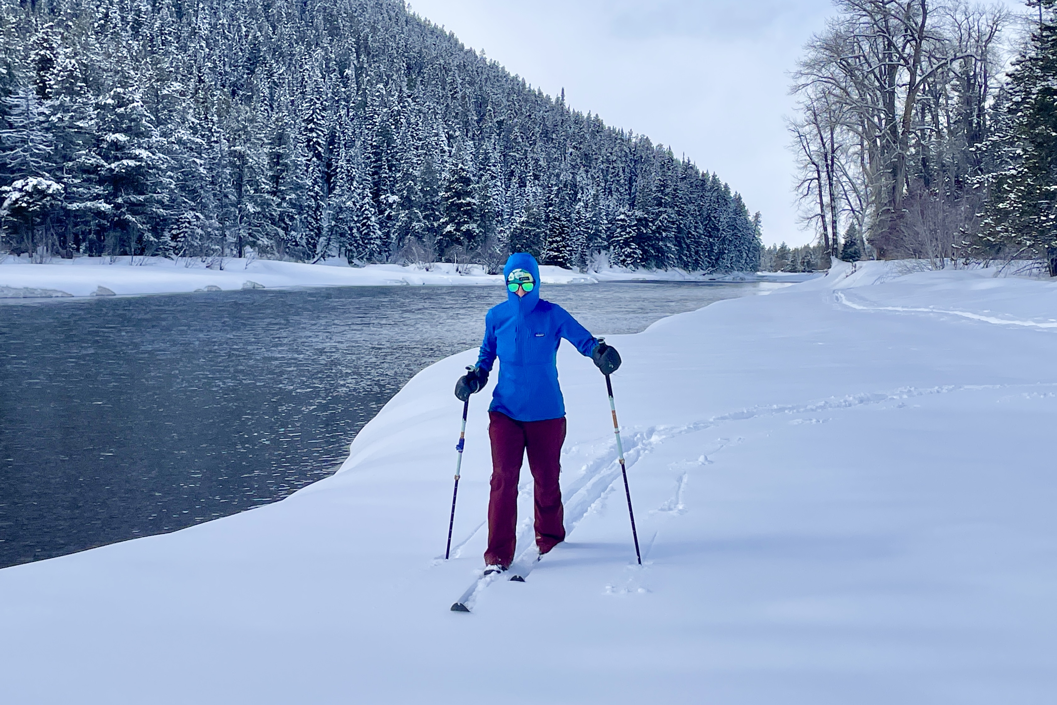 A person skis beside a calm river surrounded by snow-covered trees and mountains