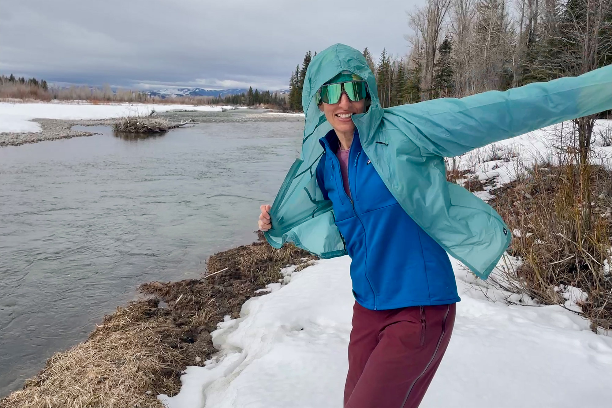 A woman stands by a snowy riverbank, smiling as she opens a light green shell jacket to show the blue Patagonia R1 Thermal Hoody underneath