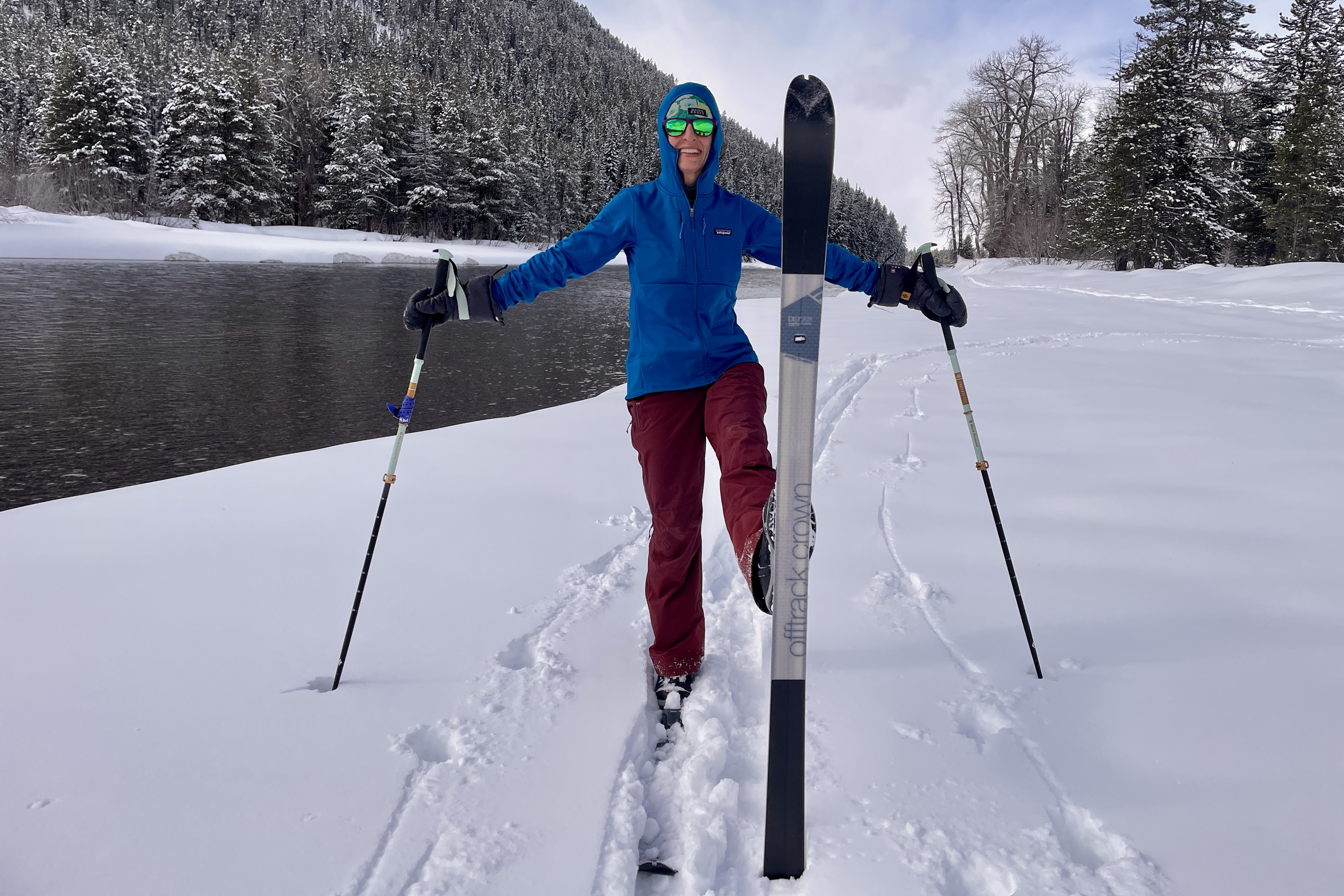 A skier in a blue Patagonia R1 Thermal Hoody and red pants stands on snowy terrain by a river, holding ski poles and balancing one ski upright in front