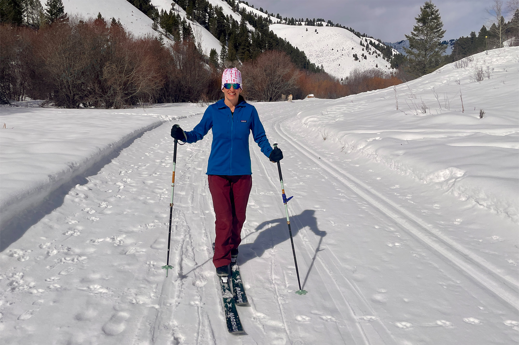 A woman skis uphill on a snow-covered trail wearing a blue jacket, maroon pants, and a pink hat