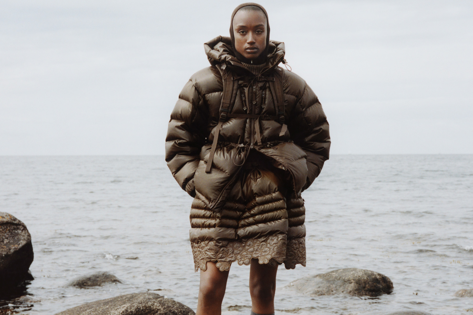 Person stands on rock in ocean wearing brown puffy clothing
