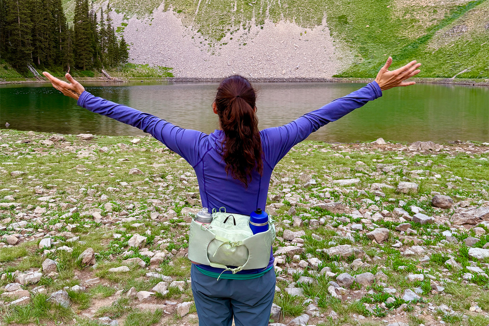 A hiker standing by a mountain lake with arms raised, wearing the Osprey Tempest & Talon 6 Waistpack
