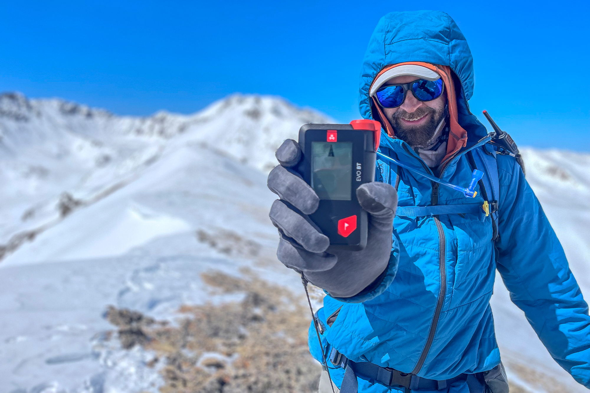 A smiling hiker holds up an avalanche beacon on a snowy mountain ridge
