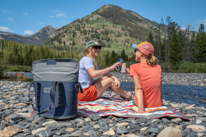 two women sitting next to river with soft cooler and drinks