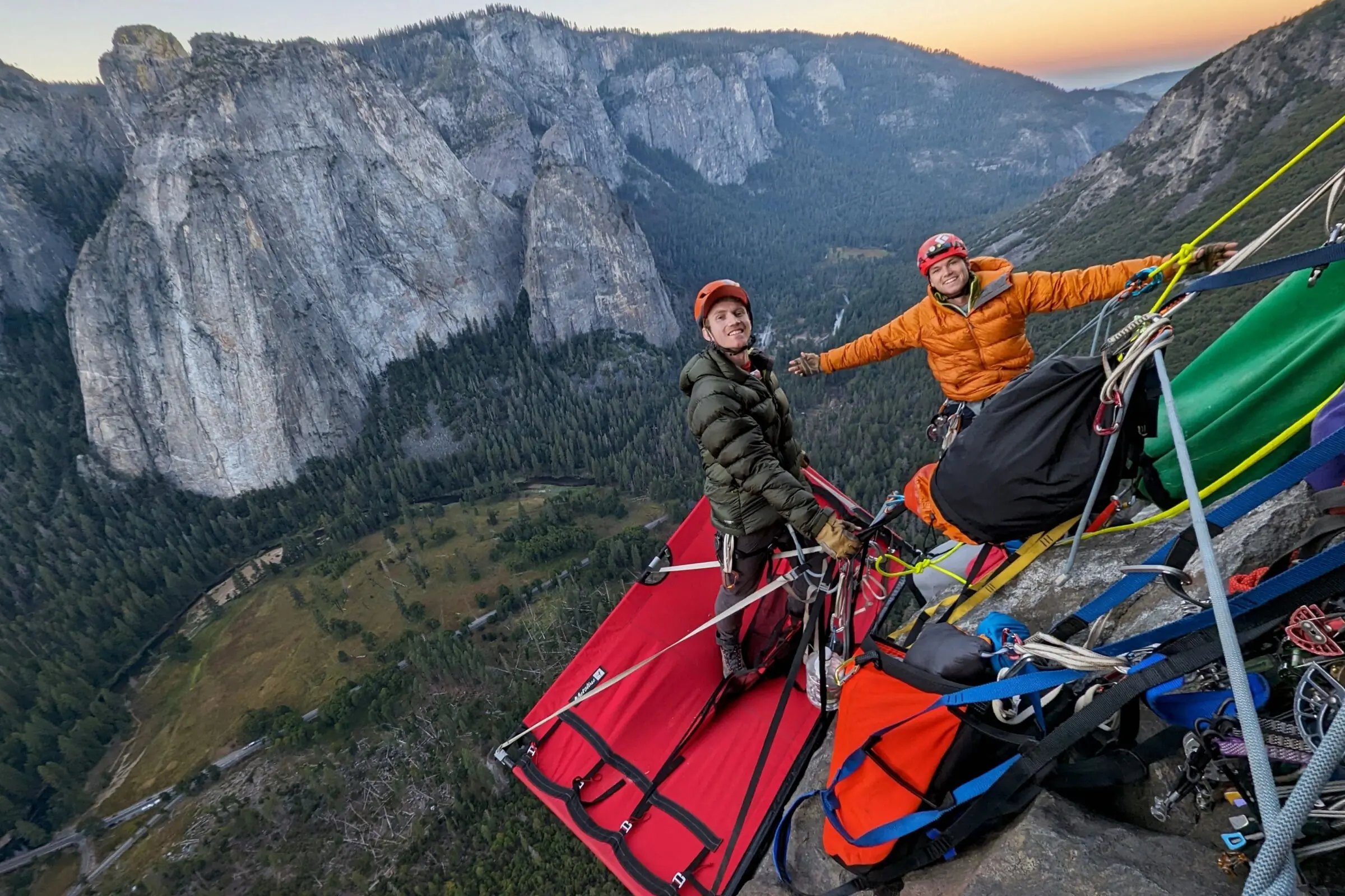 Two climbers stand on a portaledge while wearing down jackets