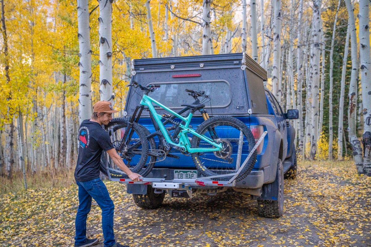 A person loading a teal mountain bike onto a 1Up heavy-duty hitch rack on a blue pickup truck