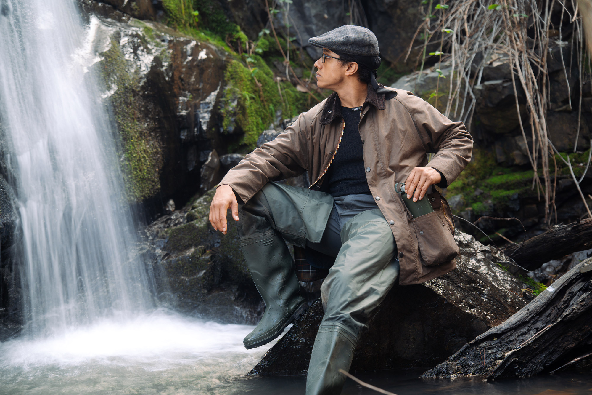 A person sits near a waterfall wearing the Barbour Beaufort Wax Jacket