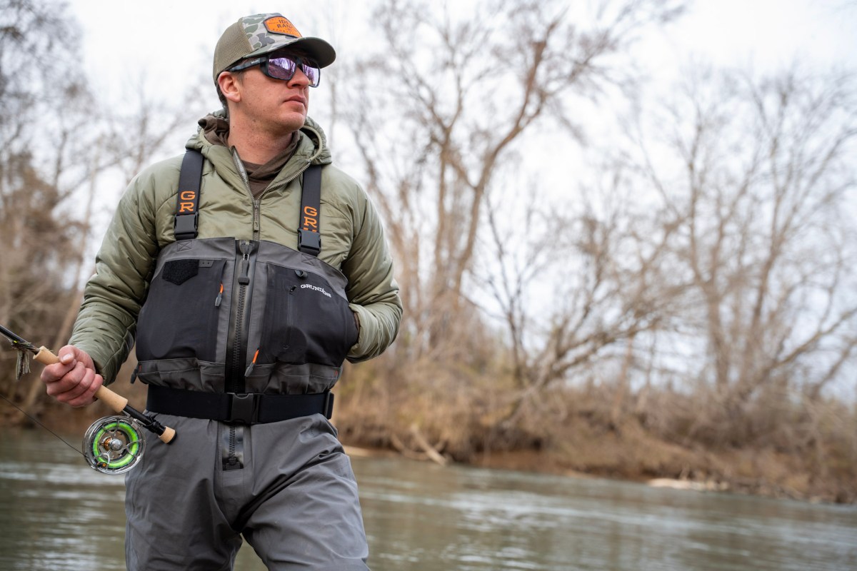 An angler in waders and sunglasses holding a fly rod in a river.