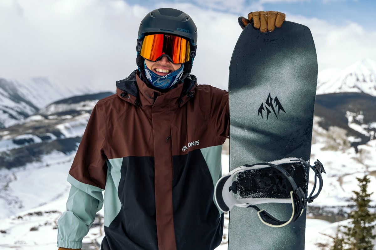 Rider holds the Jones Howler snowboard upright with mountain landscape behind