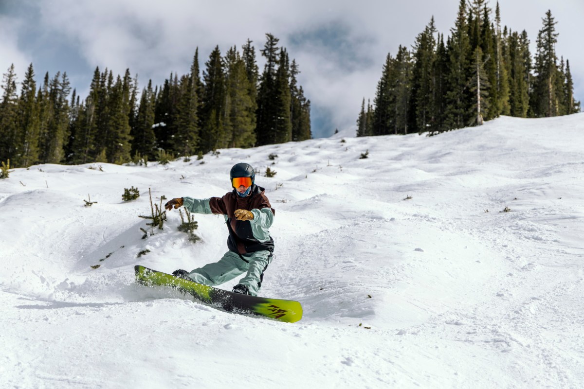 Snowboarder rides the Jones Howler through tracked snow with trees in the background