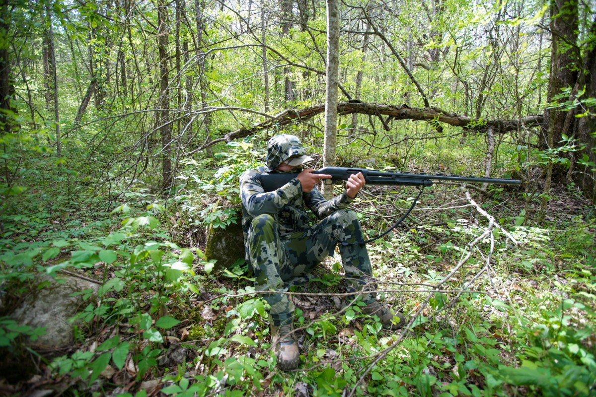 A hunter in the turkey woods sitting down, aiming a shotgun.