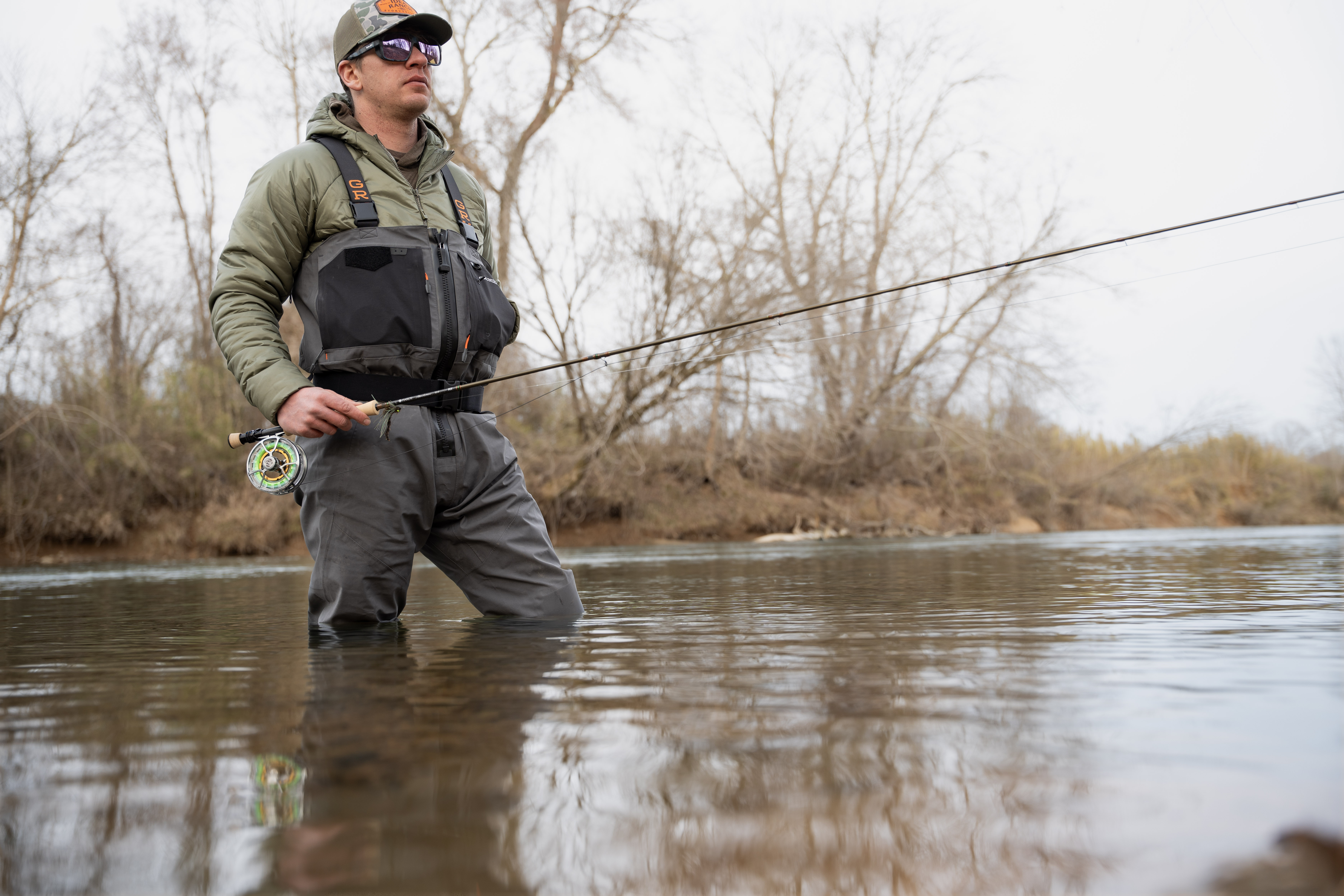 An angler wearing the Grundens Vector Zip waders standing in a river holding a fly rod.