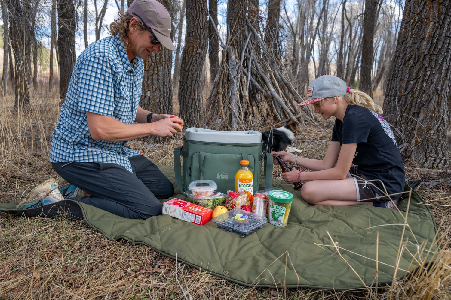 A man and child having a picnic in the woods with the REI Trail Gate cooler and snacks spread out on a blanket.