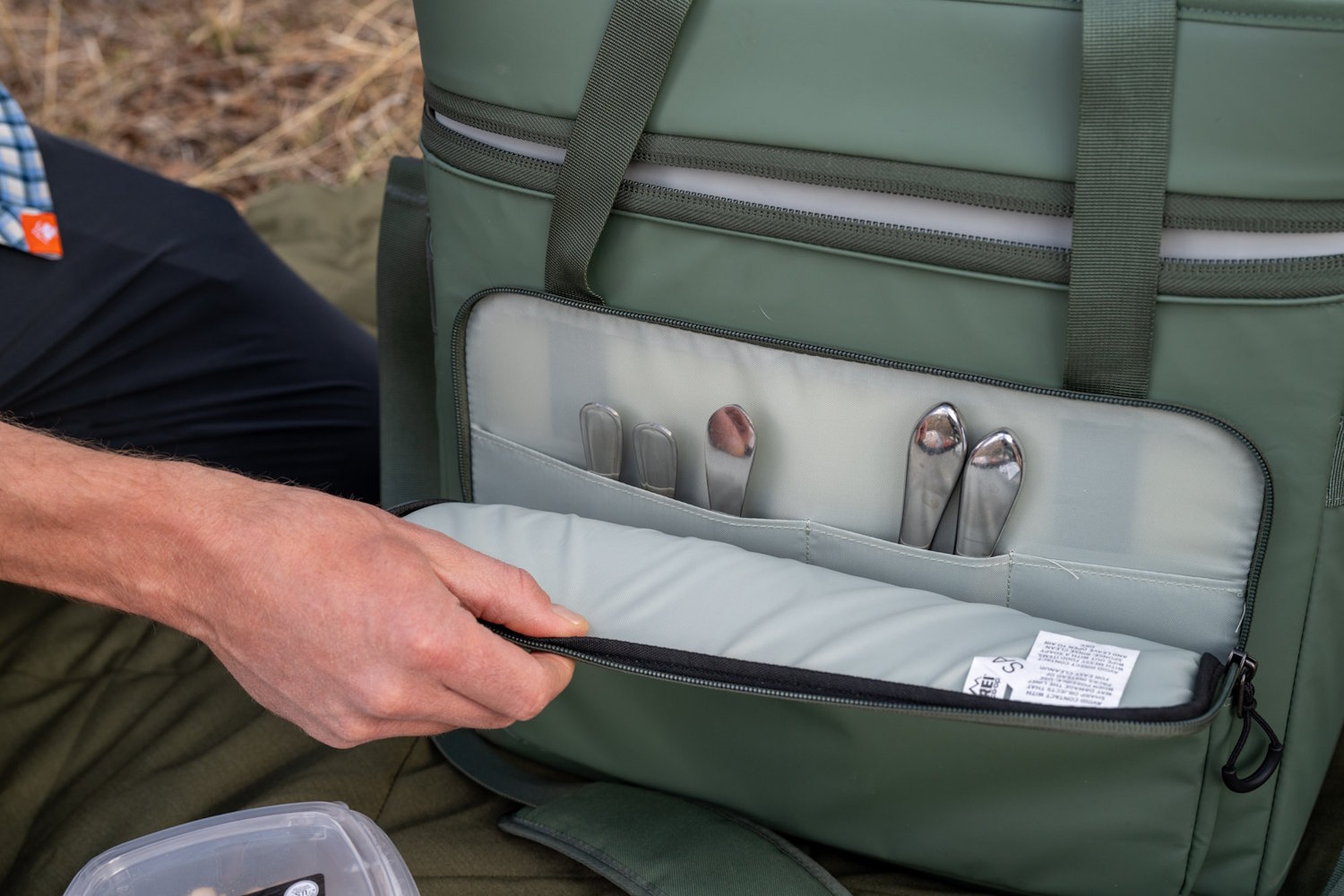 A hand opening the front zippered pocket of the REI cooler to reveal internal slots holding metal silverware.