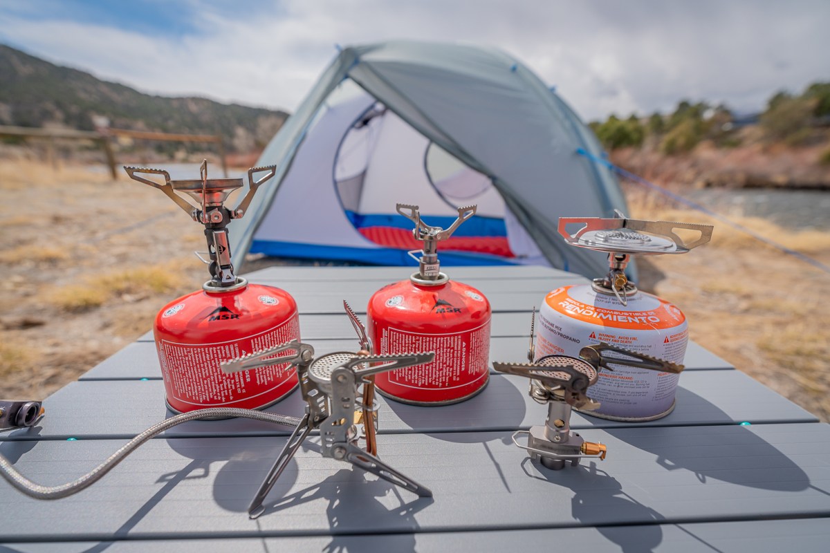 five different lightweight backpacking stoves with fuel canisters and a tent in the background