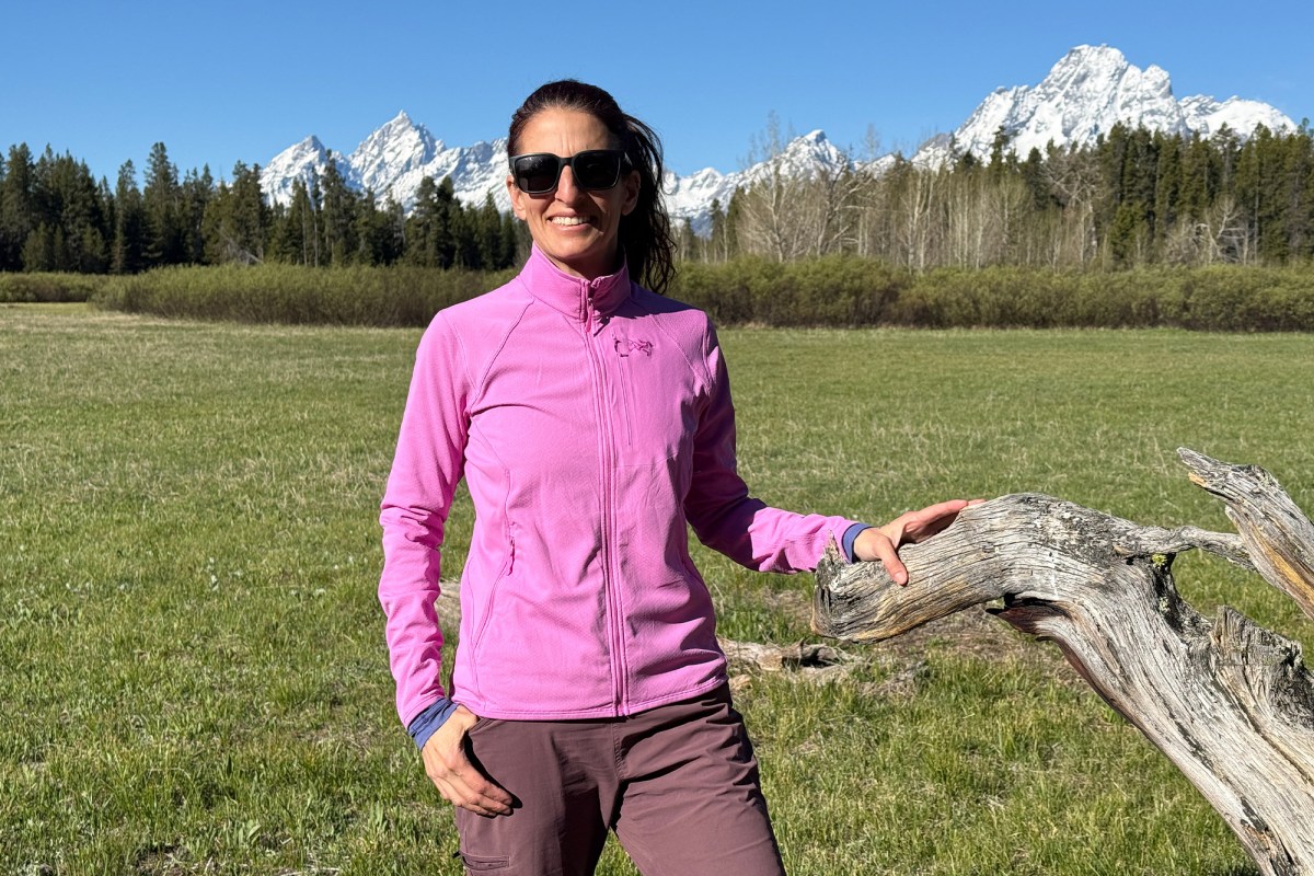 Woman wearing a pink Arc’teryx Delta Jacket in a grassy meadow with snowy mountains behind her