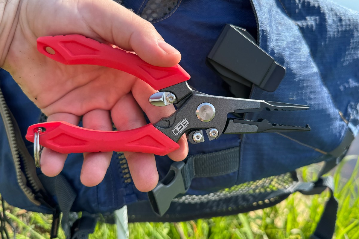 A hand holds red and black EGO fishing pliers in front of a blue life vest near the water