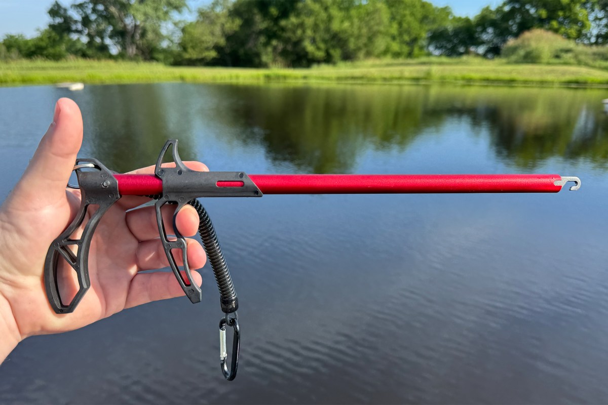 A hand holds the red and black EGO Ultra hook remover over a pond with trees and grass in the background