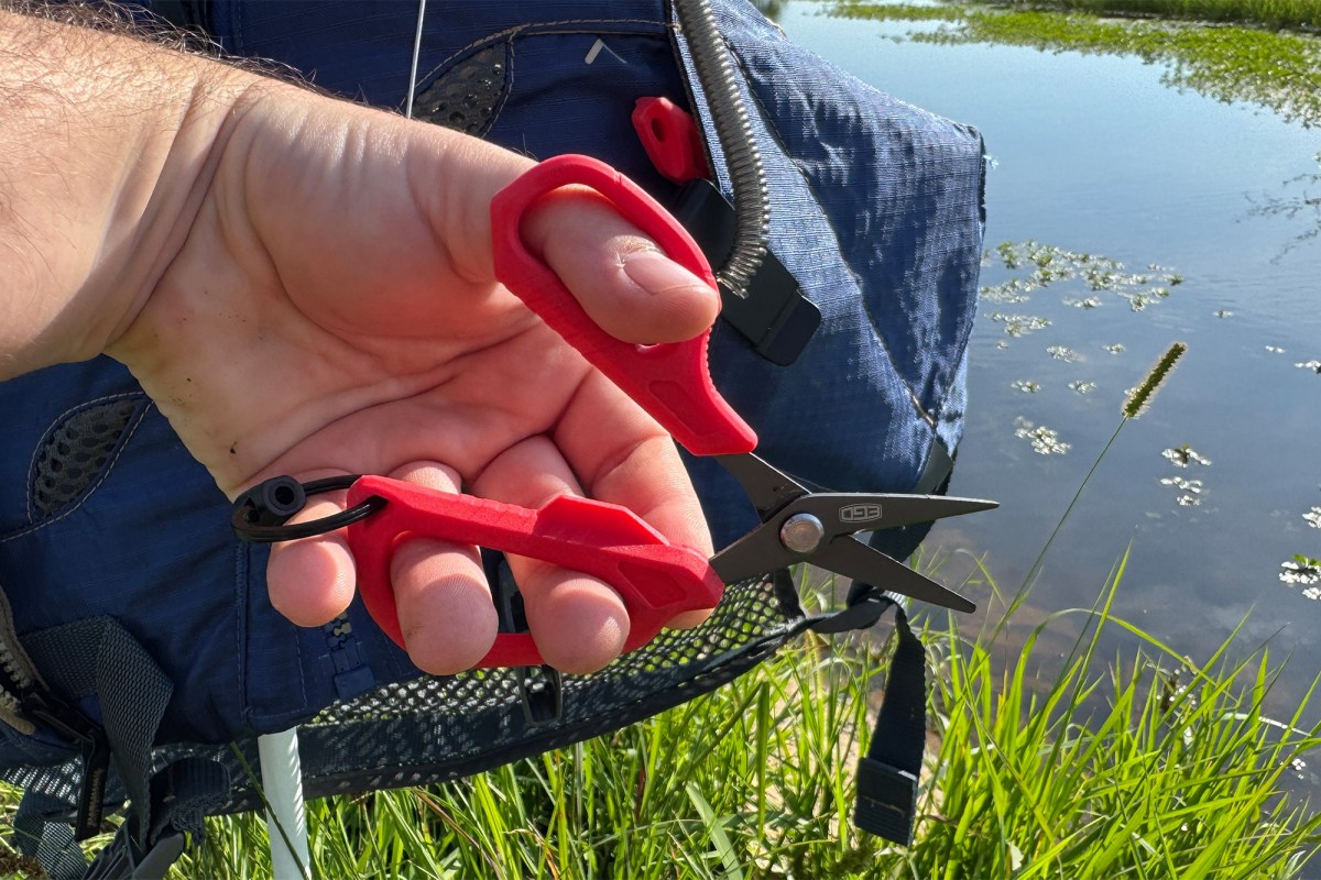 A hand holds red EGO utility scissors in front of a blue life vest and a grassy pond edge