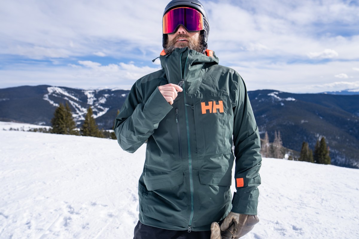 Skier adjusts the high collar on the Helly Hansen Ridge Infinity Jacket 2.0 while standing on a snowy slope