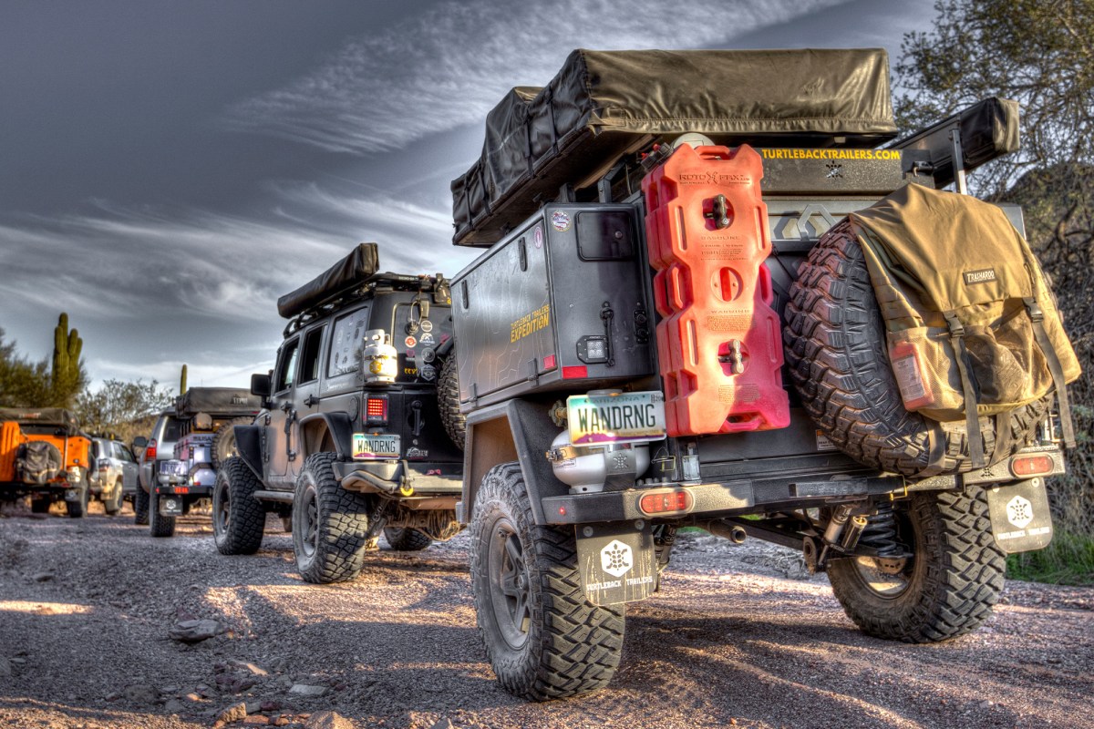 Turtleback trailer convoy with rear-mounted spare tire, fuel containers, and rooftop tent on a rocky desert road