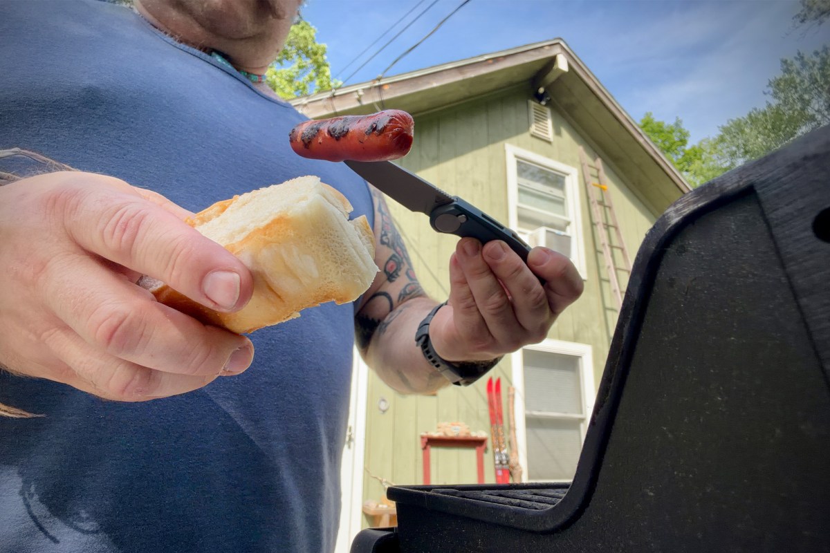 Madrone knife being used to hold a grilled hot dog beside a barbecue