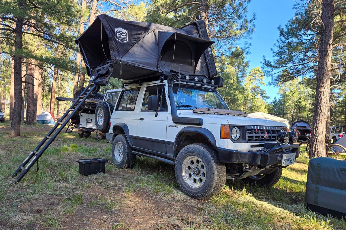White Mitsubishi Montero with a rooftop tent and ladder at a forest campsite