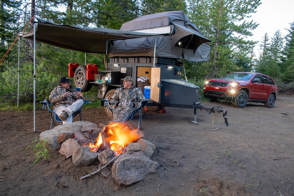 Off-road camper trailer with a rooftop tent, awning, and campfire at a forest campsite