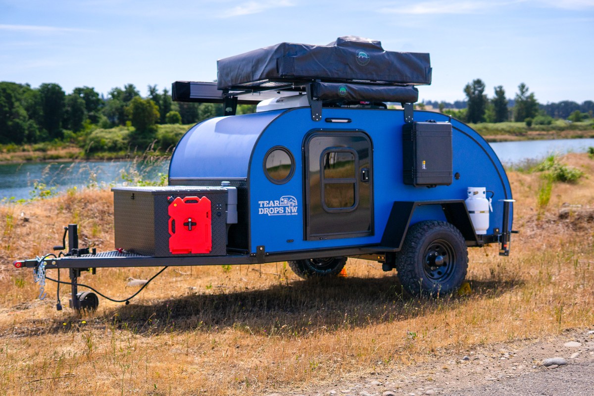 Blue teardrop camper trailer with rooftop tent, storage box, and red fuel can near a river