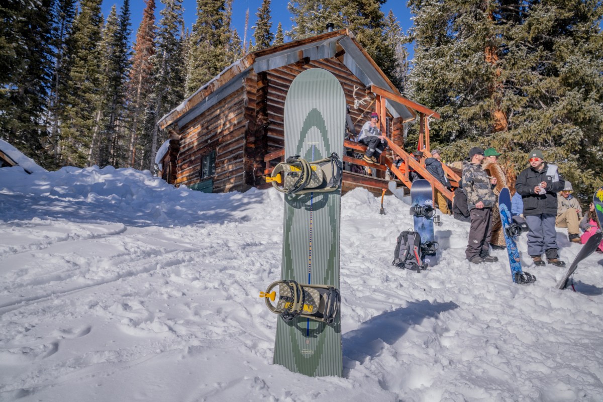 A Salomon Highpath snowboard stands upright in the snow with bindings attached near a rustic cabin and a group of snowboarders