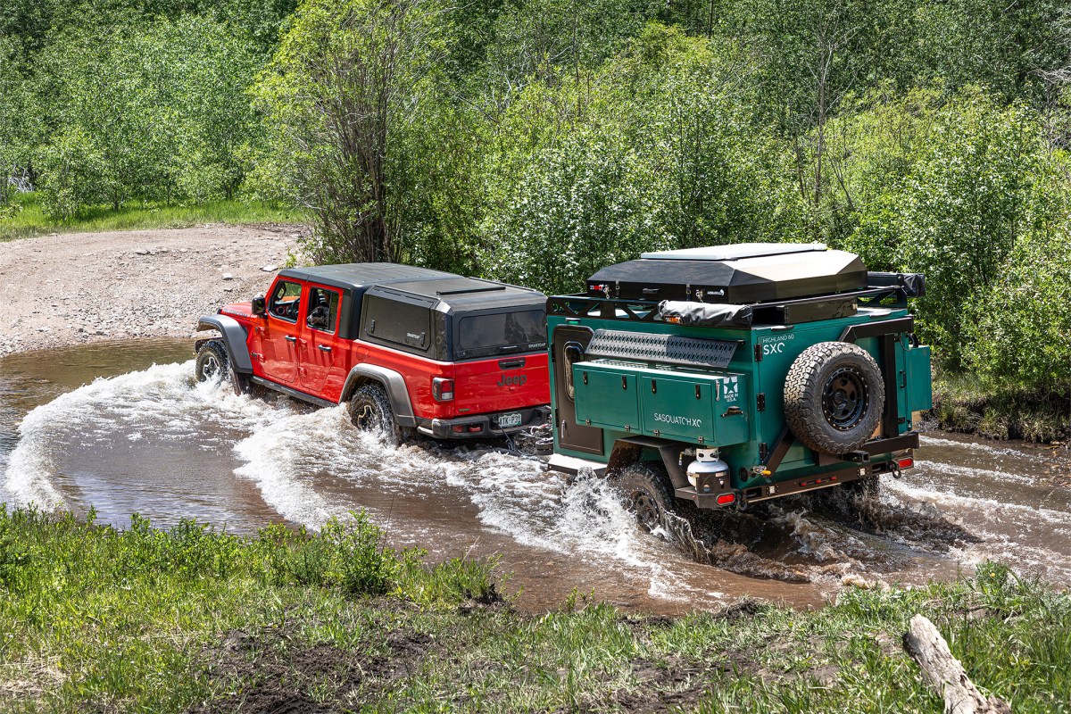 Jeep towing a green Sasquatch H60 trailer through a shallow creek