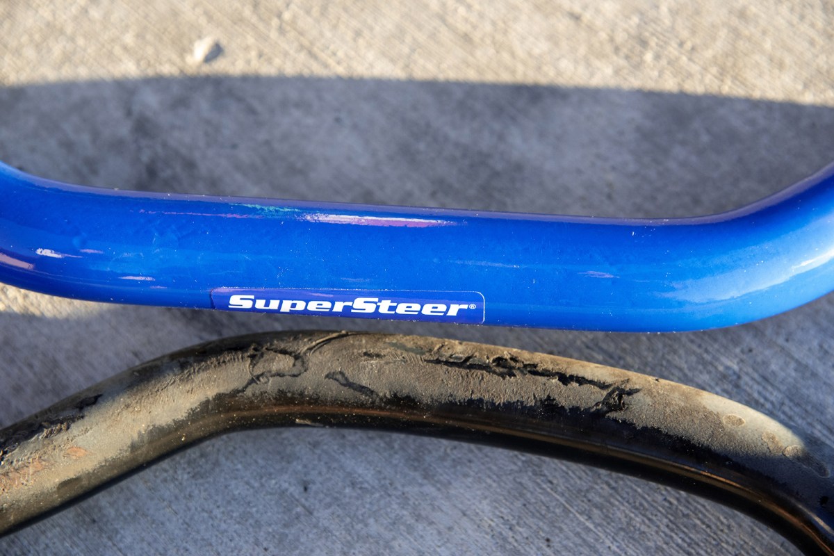 A blue SuperSteer sway bar shown next to an older black sway bar on pavement