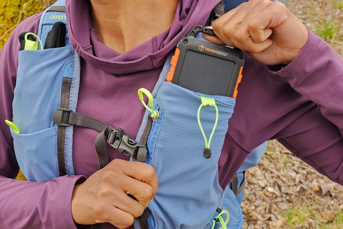 A hiker pulls a small device from the front chest pocket of a Tempest Velocity 30 backpack
