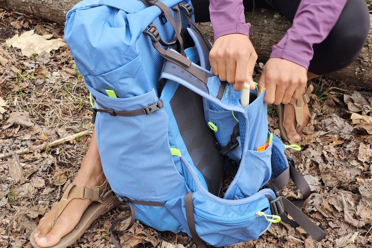 Hands place a small item into the side pocket of a blue Tempest Velocity 30 pack