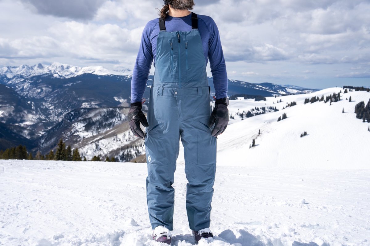 Person wearing Black Diamond Recon Bib snow pants and gloves standing on a snowy mountain landscape