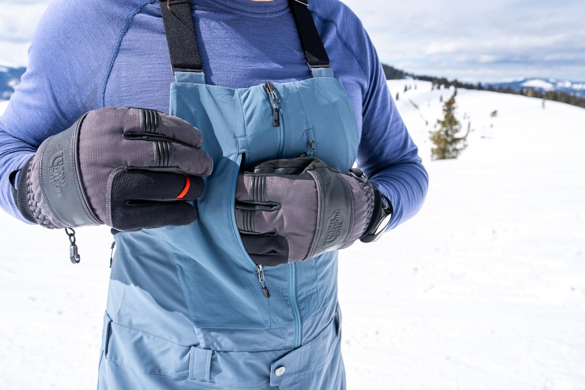 A close-up of a skier using gloved hands to access the zippered storage on the chest of the Recon bibs.