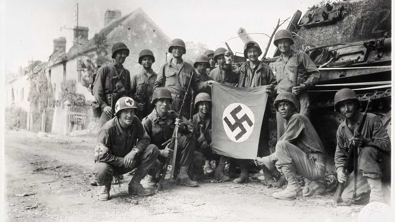 Lined_up_in_front_of_a_wrecked_German_tank_and_displaying_a_captured_swastika,_is_a_group_of_Yank_infantrymen_of_the_90th_Div._who_were_left_behind_to__mop-up__in_Chambois,_France_(49392031211)