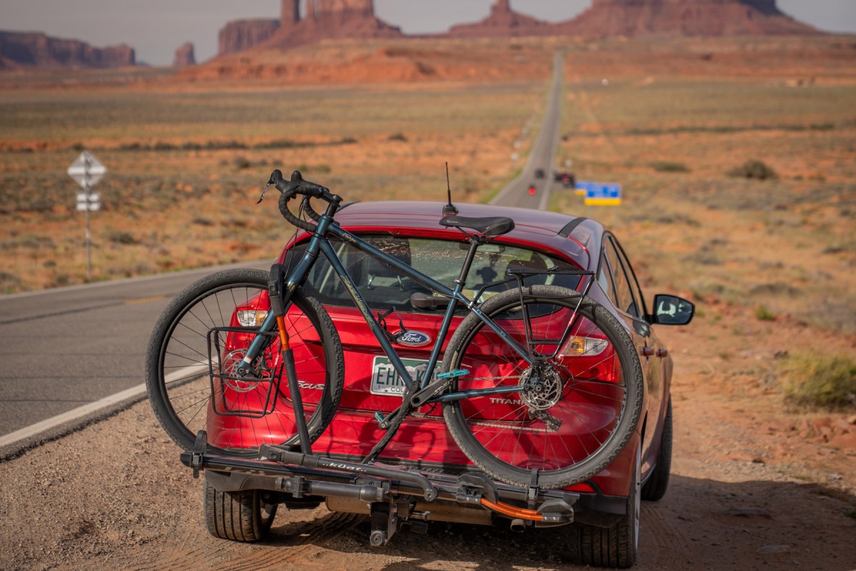 Bike secured on a Kuat NV 2.0 rack behind a red car on a desert highway