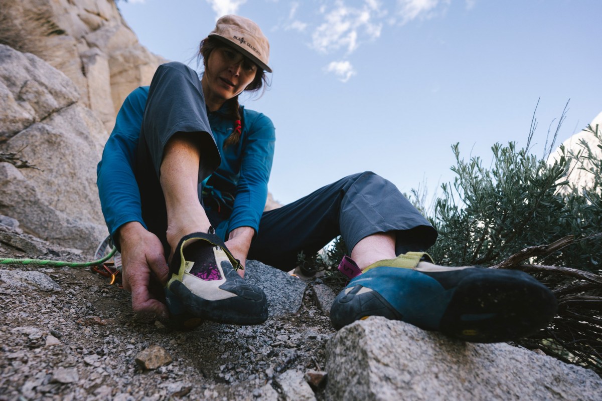 Climber putting on Skwama shoes while sitting on rocky ground before a route