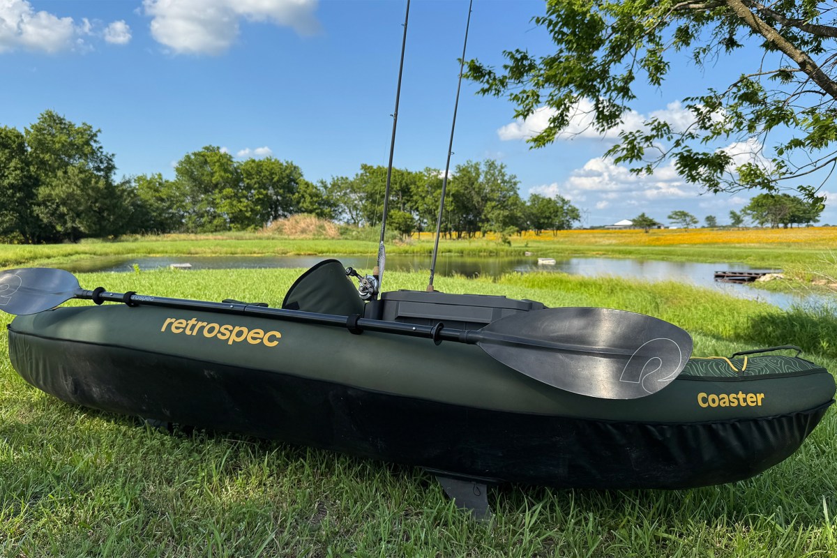 Retrospec Coaster kayak with paddle and fishing rods beside a grassy pond
