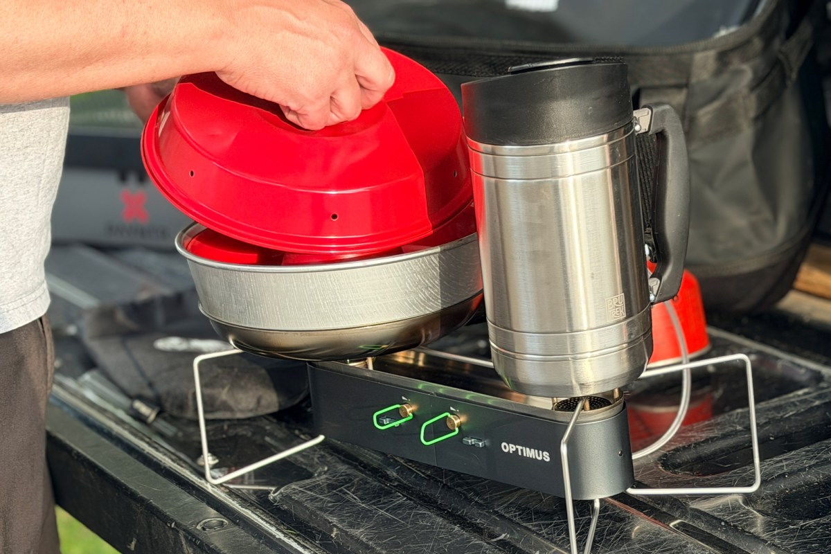 Gemini II dual-burner camp stove holds a red-lidded pot and a stainless steel mug on a truck tailgate