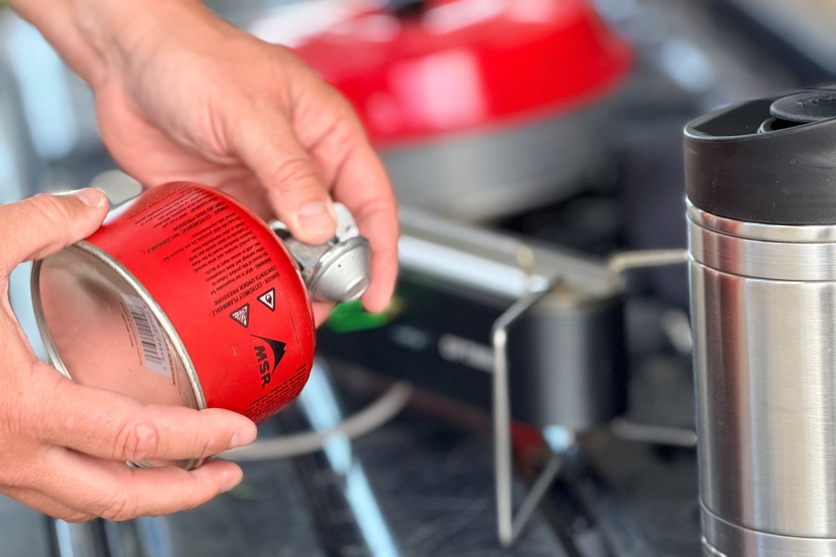 Hands attach a red fuel canister to the Optimus Gemini II dual-burner camp stove