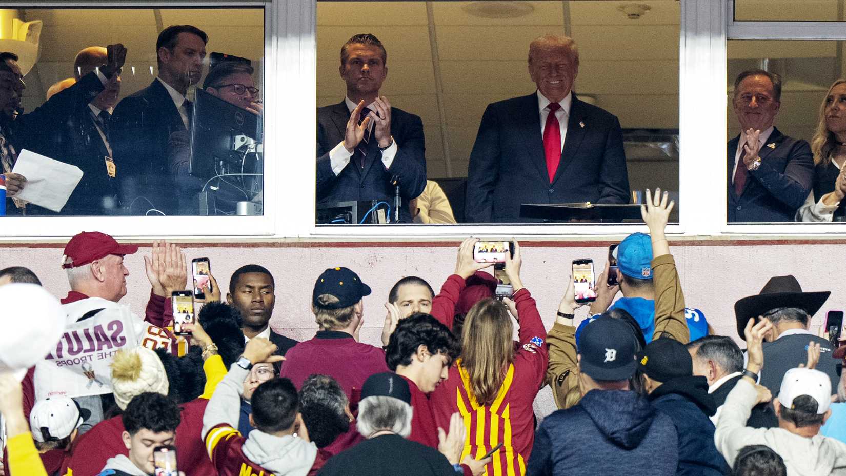 President Donald Trump stands in a stadium suite above Washington Commanders fans during an NFL Salute to Service game in Landover, Maryland.