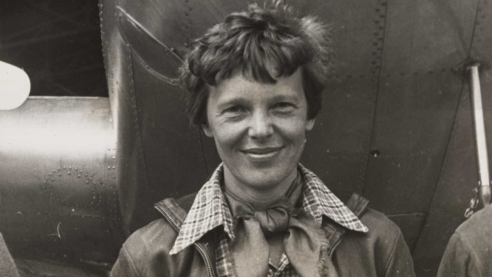 Black-and-white close-up portrait of Amelia Earhart smiling beneath the nose of her Lockheed Electra aircraft in March 1937.